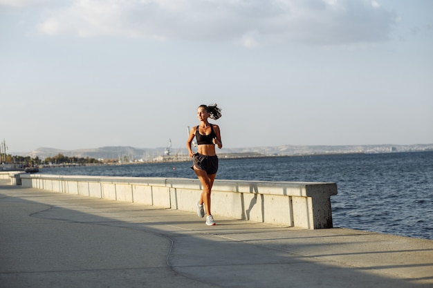 Linda garota fitness com shorts e top esportivo corre à beira-mar à beira-mar durante o pôr do sol