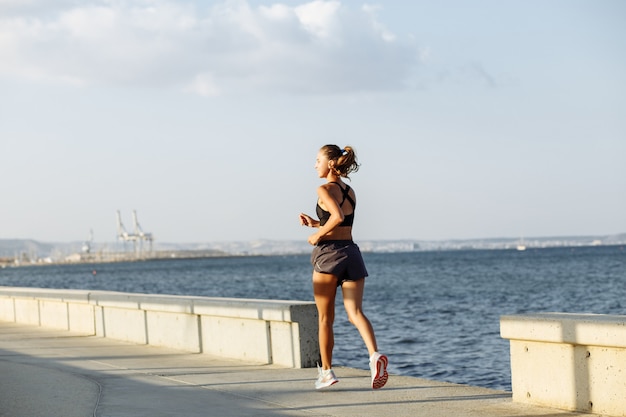 Linda garota fitness com shorts e top esportivo correndo à beira-mar à beira-mar em tempo ensolarado