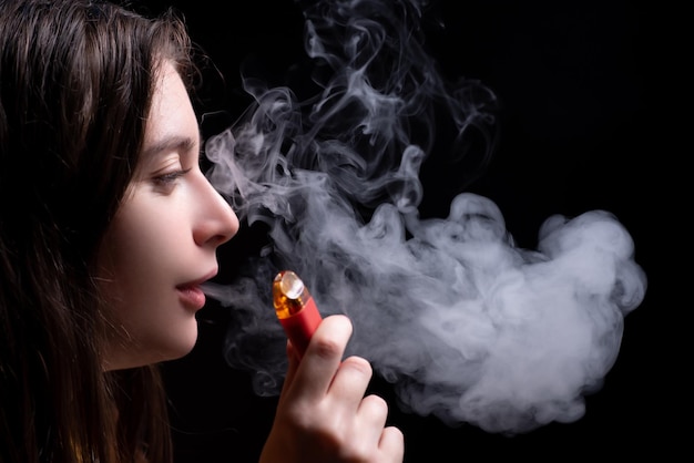 Linda garota fuma um sistema de cápsulas de vape vermelho com cartuchos substituíveis Isolado em um fundo preto O conceito de vaping de fumo moderno e nicotina