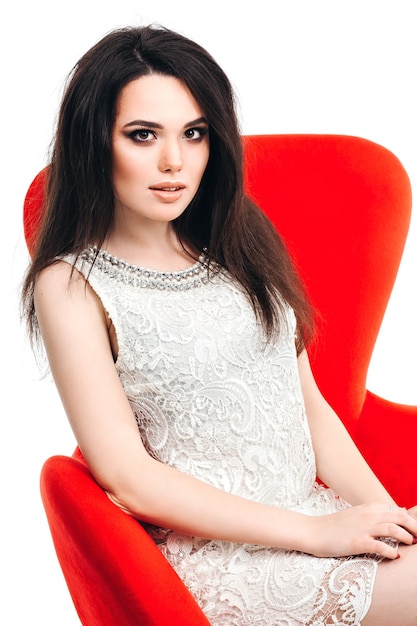 Linda garota glamorosa com uma taça de champanhe, sentada em uma cadeira vermelha isolada em um fundo branco, olhando para a câmera