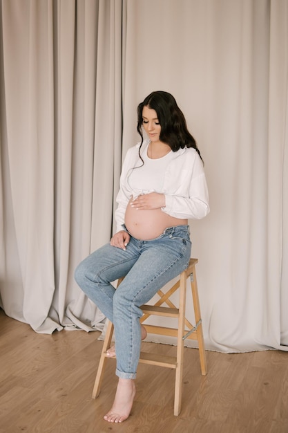 Linda garota grávida com cachos em uma camisa branca e jeans em um estúdio brilhante com um interior elegante o conceito de uma feliz gravidez e família