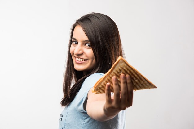 Linda garota indiana asiática comendo sanduíche grelhado, em pé, isolada sobre um fundo amarelo ou branco