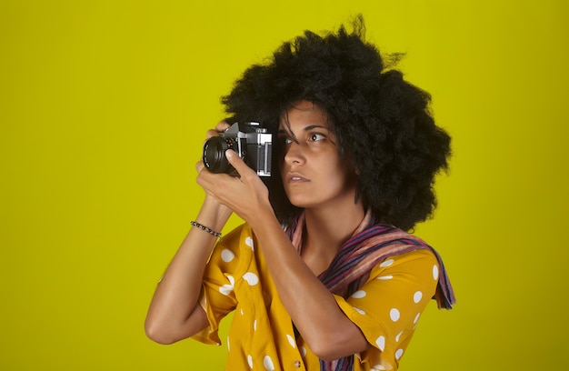 Linda garota indiana com um penteado afro encaracolado falando fotos com a câmera retro na parede amarela