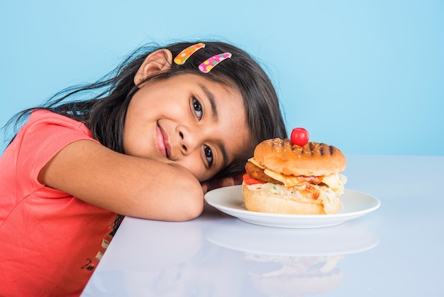 Linda garota indiana ou asiática comendo hambúrguer saboroso, sanduíche ou pizza em um prato ou caixa. Permanente isolado sobre fundo azul ou amarelo.