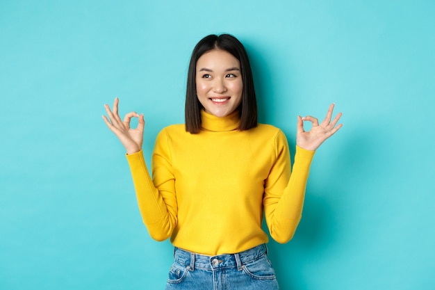 Linda garota japonesa com um suéter amarelo, mostrando sinais de ok e sorrindo, olhando para a esquerda e o logotipo