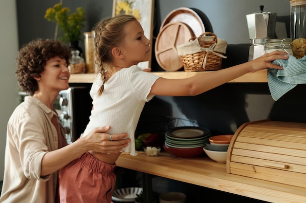 Linda garota jovem com espanador limpando prateleiras de madeira com utensílios de cozinha