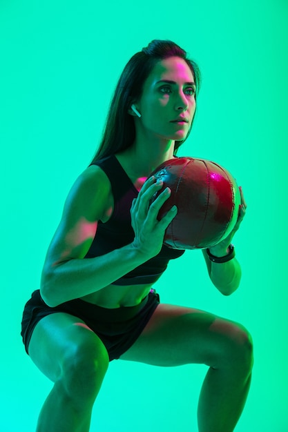 Linda garota jovem e confiante fitness isolada na parede de néon verde, usando fones de ouvido sem fio, malhando com uma medicine ball, agachada