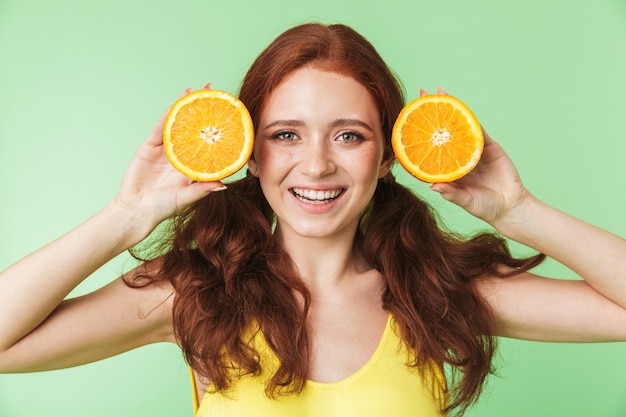 linda garota jovem ruiva animada posando isolado sobre o fundo da parede verde com frutas de vitaminas cítricas.