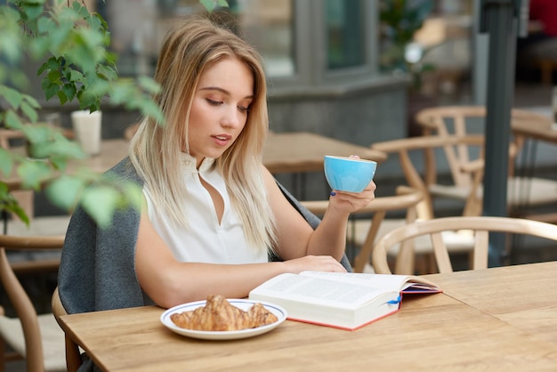 Linda garota lendo livro, bebendo café e comendo croissant ao ar livre