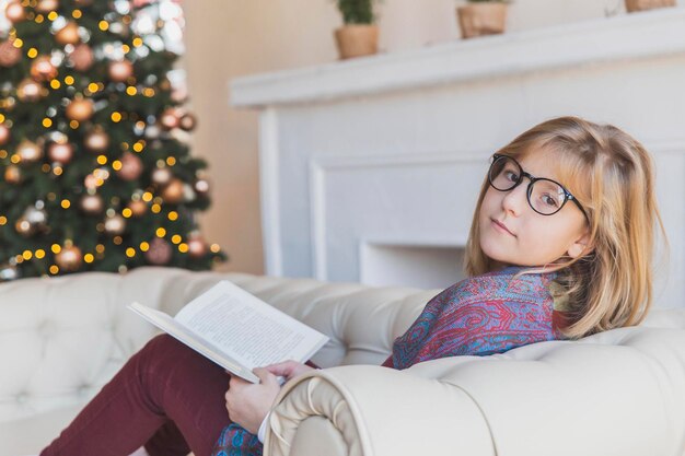 Linda garota lendo um livro no sofá perto da árvore de natal