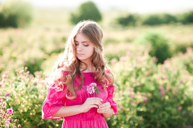 Linda garota loira adolescente usando um vestido rosa segurando uma flor rosa ao ar livre