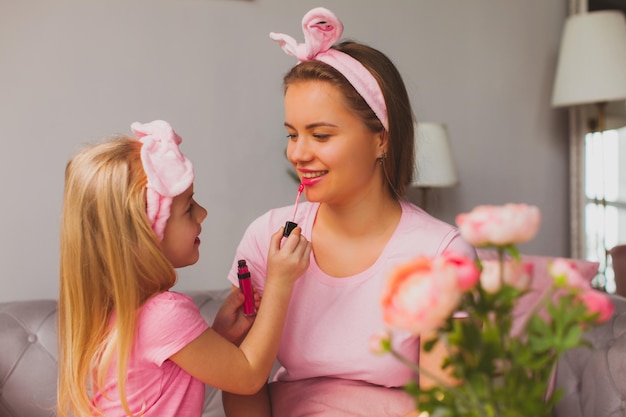 Linda garota loira aplicando brilho rosa nos lábios de sua mãe Mulher alegre e garota jogando salão de beleza em casa