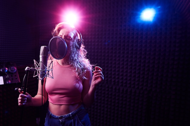 Linda garota loira cantando emocionalmente em um estúdio de gravação com microfone e fones de ouvido profissionais, cria um novo álbum de faixas, artista vocal em luz de néon azul rosa