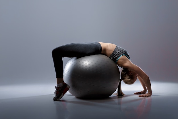 linda garota loira com roupas esportivas, sentada em uma bola de fitness em um fundo escuro