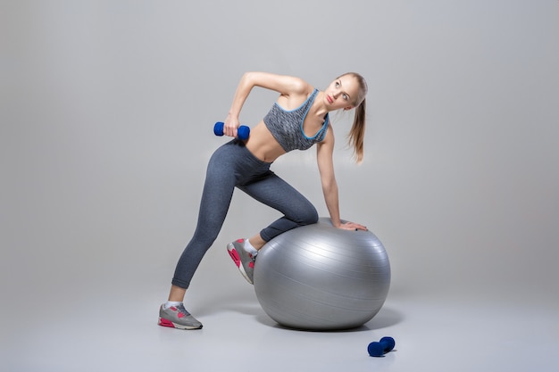 linda garota loira em roupas esportivas faz exercícios com halteres na bola de fitness em um fundo cinza