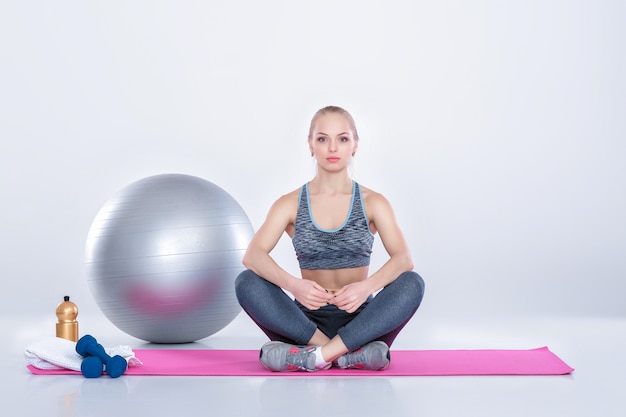 Linda garota loira em roupas esportivas fazendo exercícios em um tapete de fitness em um fundo cinza