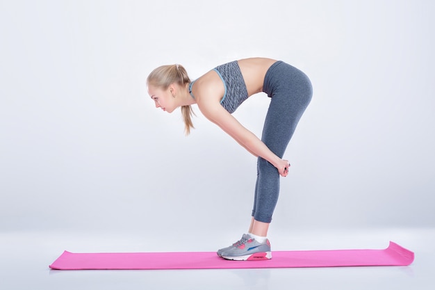 linda garota loira em roupas esportivas fazendo exercícios em um tapete de fitness em um fundo cinza