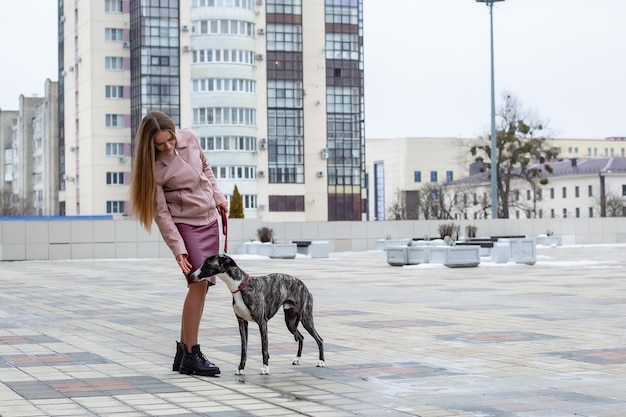 linda garota loira mantém um cachorro whippet na coleira na cidade