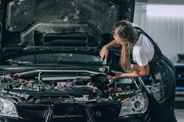 Linda garota mecânica em um macacão preto e uma camiseta branca muda o óleo em um carro preto. conceito de conserto de carro