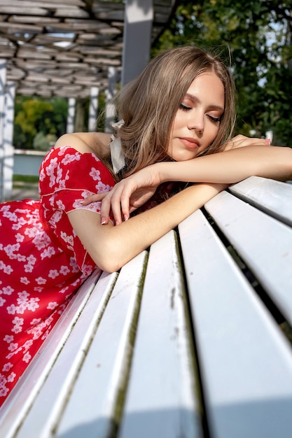 Linda garota, modelo, mulher de cabelos castanhos com cabelos longos em um vestido vermelho, senta-se num banco e posa com um olhar descontraído e lânguido.