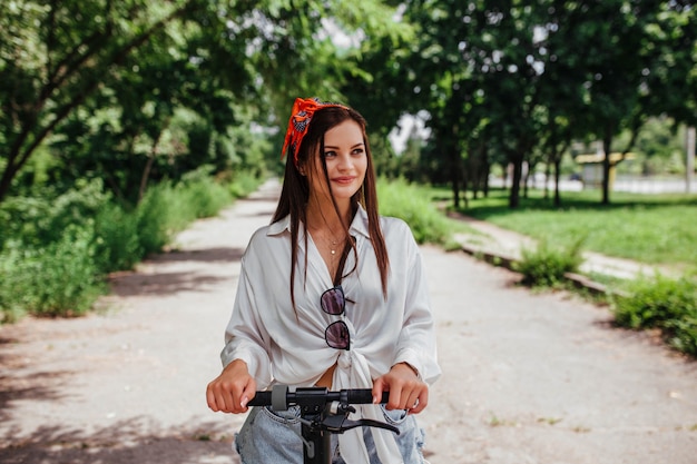 Linda garota morena anda em uma scooter de eletrodo no parque, ela está vestindo uma camisa branca. conceito de eco transporte e aluguel.