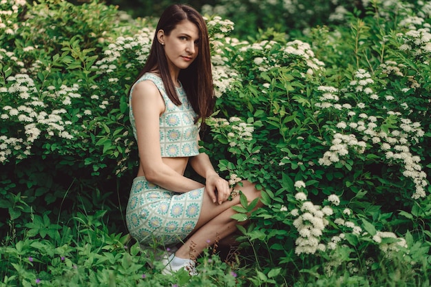 Linda garota morena senta-se ao lado do arbusto de flores na floresta e relaxando em um dia ensolarado e quente.