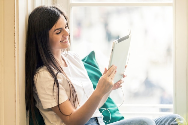 Linda garota morena sorridente usa camiseta branca usando computador tablet para videochat sentado no parapeito