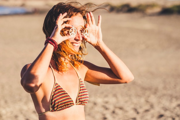 Linda garota mostrando o símbolo da paz nos olhos dela segurando as mãos - mulher de biquíni na praia sorrindo e olhando para o camer com o sol no rosto