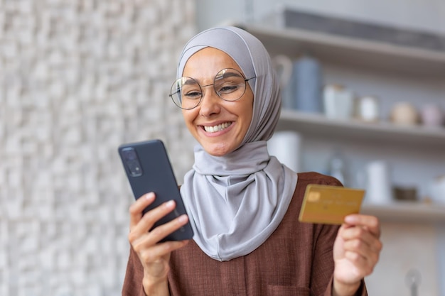 Linda garota muçulmana se aproxima da mulher da cozinha em hijab sorrindo e feliz segurando smartphone e