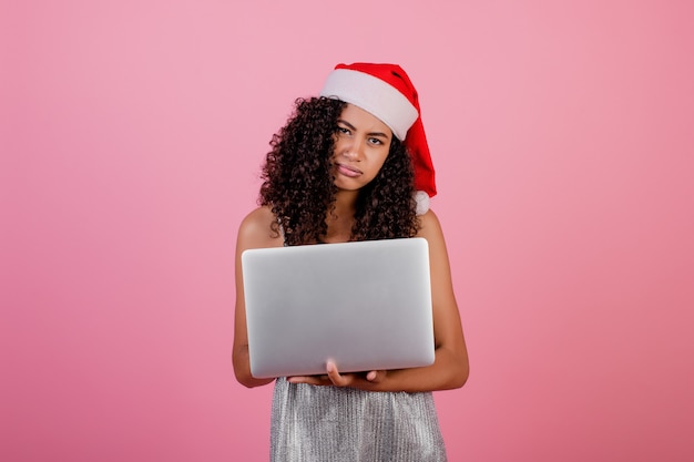 Linda garota negra com laptop usando chapéu de natal de férias e vestido isolado sobre rosa
