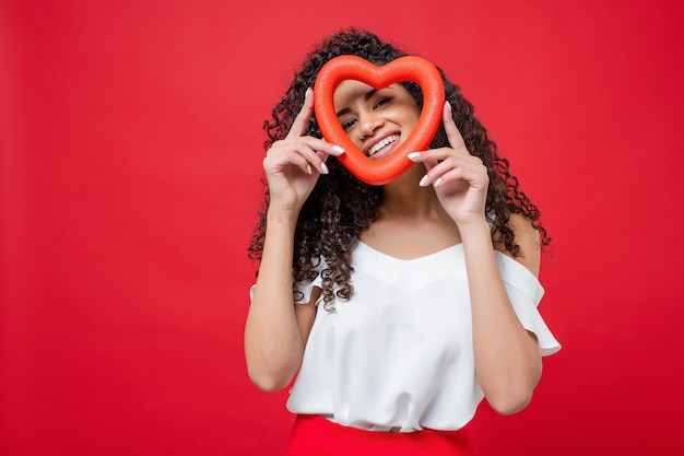 Linda garota negra, olhando através da forma do coração com sorriso feliz isolado no vermelho