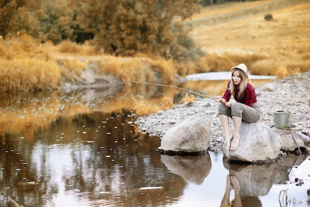 Linda garota no outono à beira do rio com uma vara de pescar