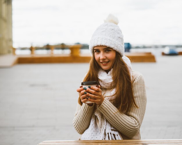 Linda garota, olhando para a câmera, segurando café, chá em caneca de plástico reutilizável no outono