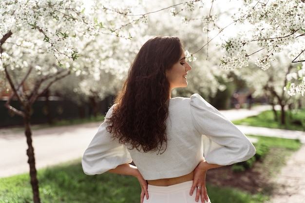 Linda garota perto de árvores florescendo no parque. caminhada ao ar livre em dia de sol. estilo de vida de uma jovem.