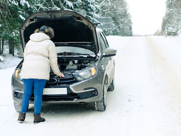 Linda garota perto do motor de um carro quebrado em uma estrada de neve de inverno. congelou e pede ajuda