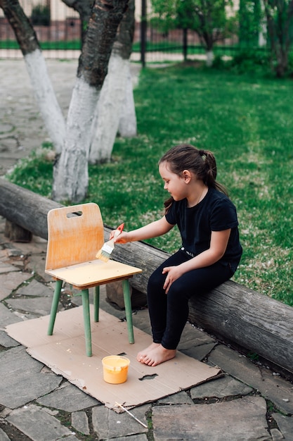 Linda garota pinta uma cadeira de madeira com tinta amarela, um hobby ecológico infantil