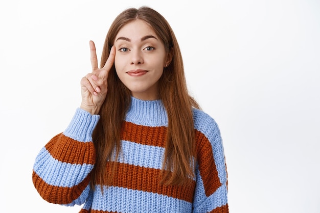 Linda garota positiva mostrando paz de vsign perto do rosto sorrindo e parecendo feliz com a câmera parada no suéter contra o fundo branco