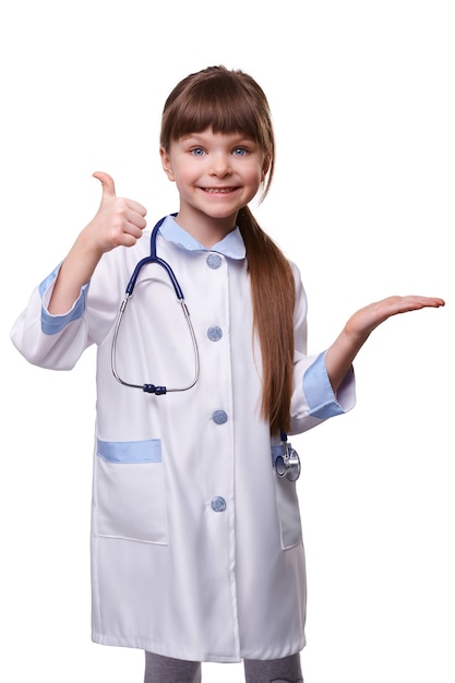 Linda garota pré-escolar sorridente, vestindo um jaleco médico com estetoscópio, mostrando o gesto do polegar e segurando a publicidade do produto na palma da mão