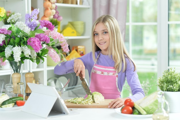 Linda garota preparando salada fresca na mesa da cozinha com tablet em casa