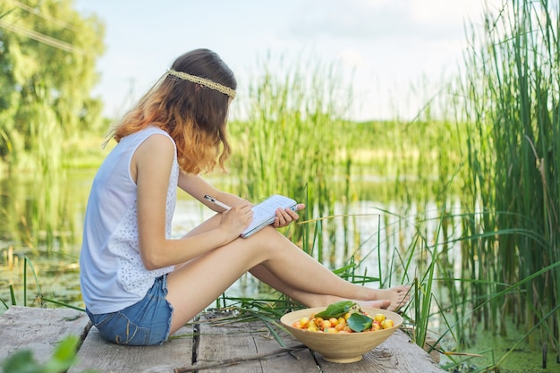 Linda garota romântica adolescente sentada na ponte perto da água, a garota escreve no caderno experiências pessoais, tigela de cerejas, apreciando a beleza da natureza pitoresca e as férias de verão, copie o espaço