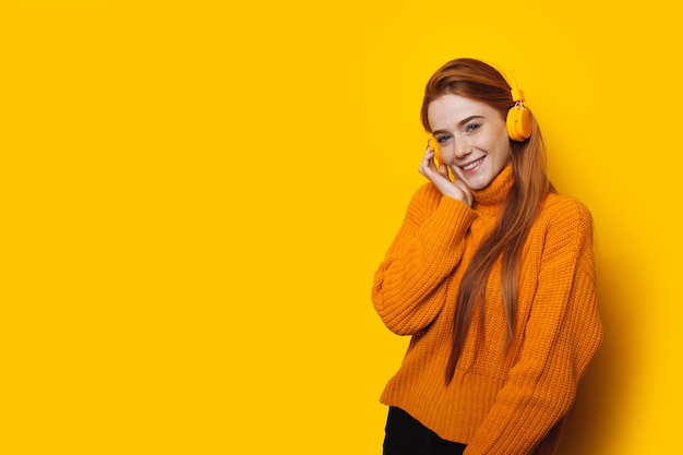 Linda garota ruiva caucasiana com sardas usando fones de ouvido posando em uma parede amarela com espaço livre