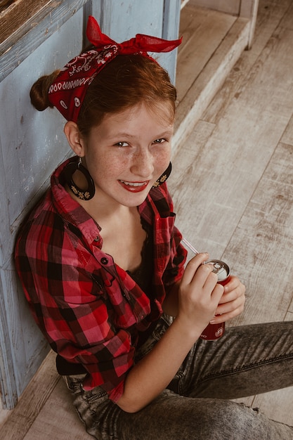 Linda garota ruiva com sardas, vestida com uma bandana vermelha e uma camisa quadriculada e bebendo um refrigerante