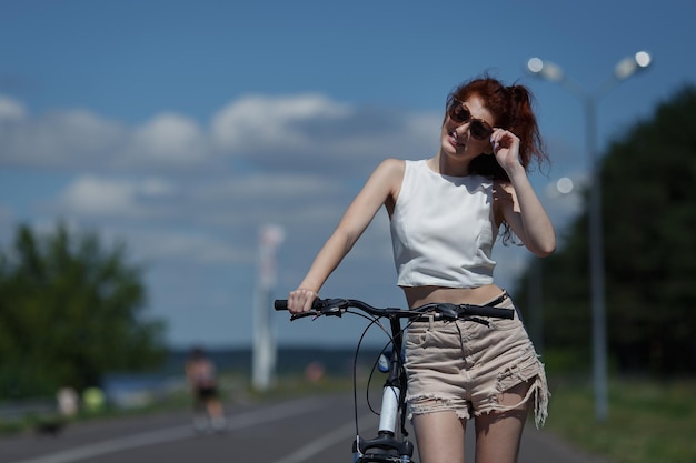 Linda garota ruiva de shorts e tops posando em pé com uma bicicleta em um dia quente de sol