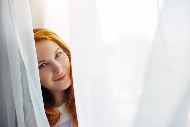 Foto linda garota ruiva jovem olha para fora da cortina branca, close-up, espaço de cópia. retrato de uma bela mulher sorridente com uma pele perfeita sobre um fundo claro e desfocado.