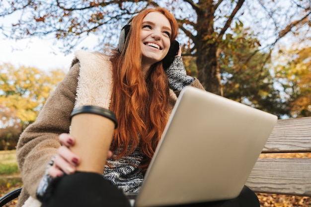 Linda garota ruiva ouvindo música com headpones enquanto está sentada em um banco, usando um laptop