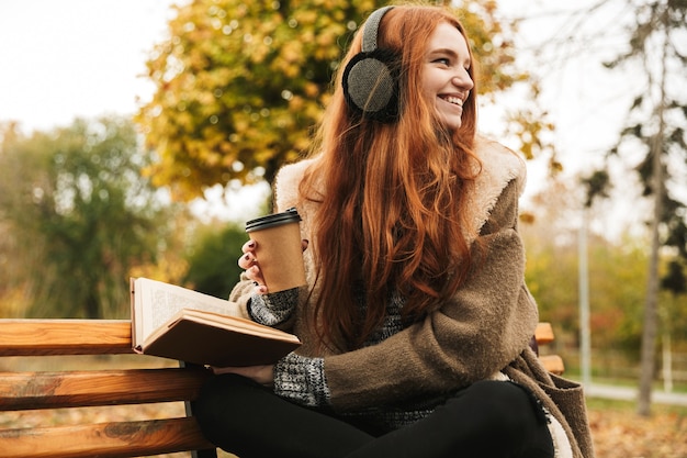 Linda garota ruiva ouvindo música com pontas de cabeça enquanto está sentada em um banco, lendo um livro, bebendo café