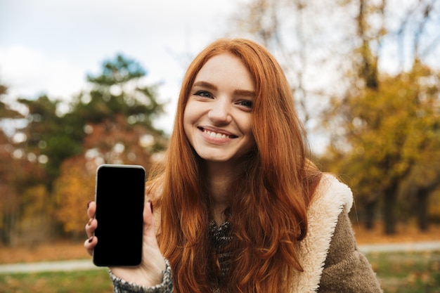 Linda garota ruiva ouvindo música enquanto está sentada em um banco, usando o telefone celular, mostrando a tela do celular