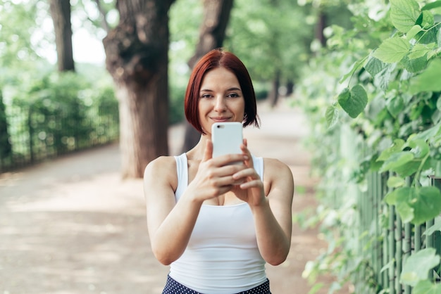 Linda garota ruiva segurando um smartphone com faz uma foto enquanto caminhava no parque.