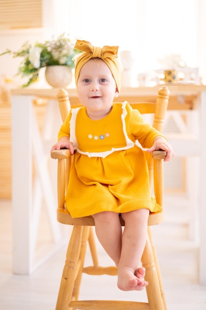 Linda garota saudável em vestido de algodão amarelo sentado em uma cadeira alta na cozinha da casa alimentando crianças com comida de bebê