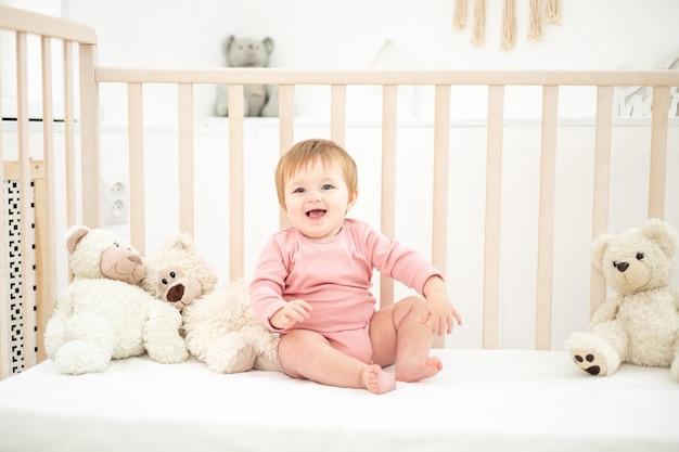 Linda garota saudável sentada em um berço com ursinhos de pelúcia na roupa de cama branca no quarto em casa bebê dormindo em seu berço